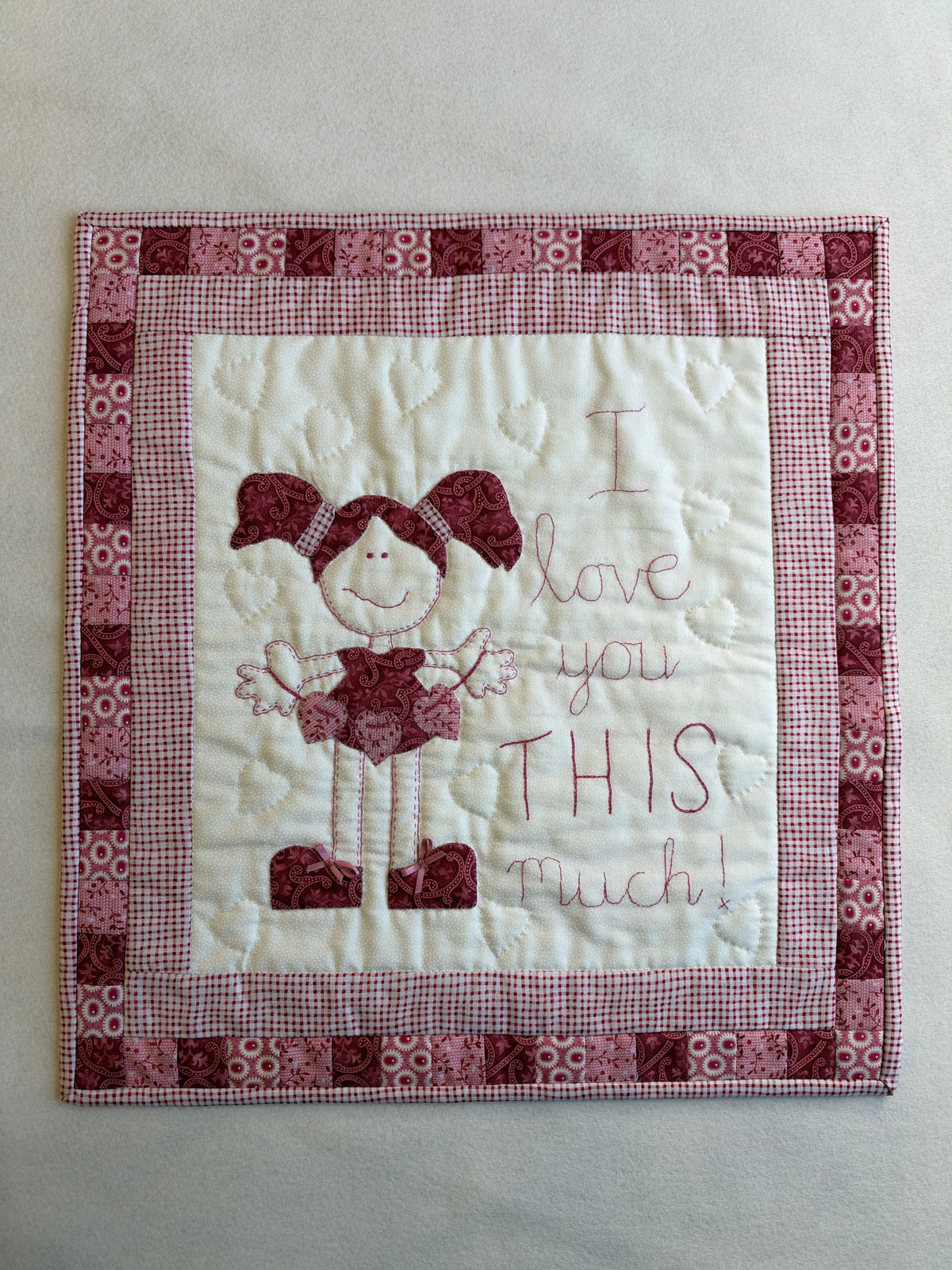 The I Love You This Much wall hanging remade in pinks and reds: a girl with pigtails beside the embroidered words