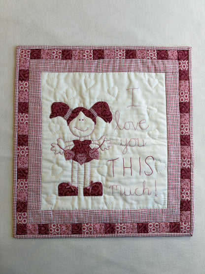The I Love You This Much wall hanging remade in pinks and reds: a girl with pigtails beside the embroidered words