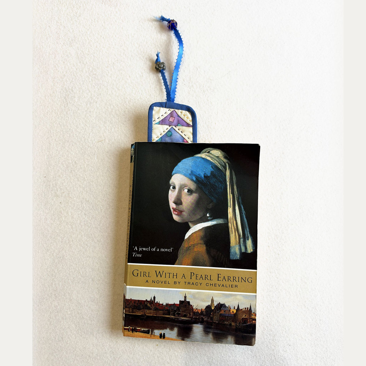 The blue Flying Geese Bookmark tucked into a paperback, with its ribbon and beads showing at the top