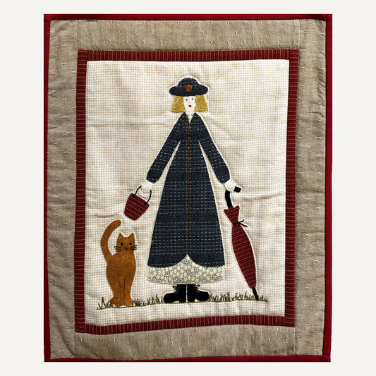 Miss Marigold wall hanging: a lady in a navy coat and hat with a basket, an umbrella and a ginger cat, framed in red and taupe borders