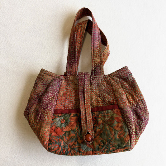 Pippa Bag made up in rust and purple batiks with a green and orange pocket, a looped strap and a wooden toggle fastening